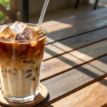Café gelado refrescante com cubos de gelo e um toque de leite, servido em um copo com canudo sobre uma mesa de madeira sob luz solar.