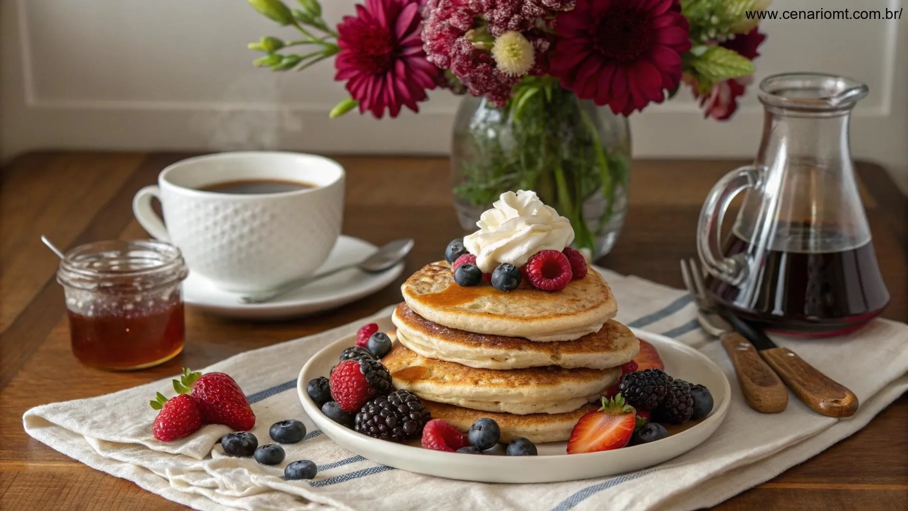 Mesa de café da manhã aconchegante com uma pilha de panquecas e várias coberturas como frutas frescas, chantilly e xarope de bordo, acompanhada por uma xícara de café e um vaso com flores.
