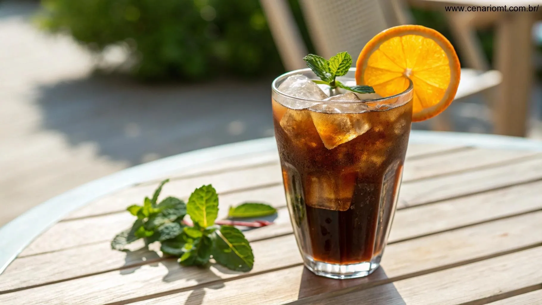 Café gelado servido em um copo transparente com uma fatia de laranja e folhas de hortelã, em uma mesa ao ar livre ao sol.