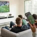 Pessoas assistindo um jogo de futebol entre Rayo Vallecano e Real Valladolid em uma televisão e celular, com foco em estatísticas ao vivo.